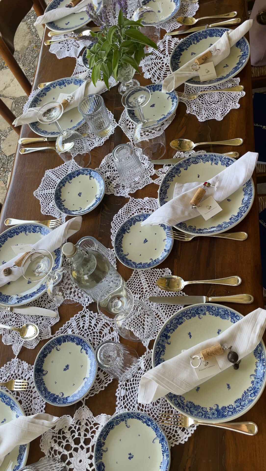 A table with blue plates and glasses and cutlery. On the wodden table under the plates are white Crochet items