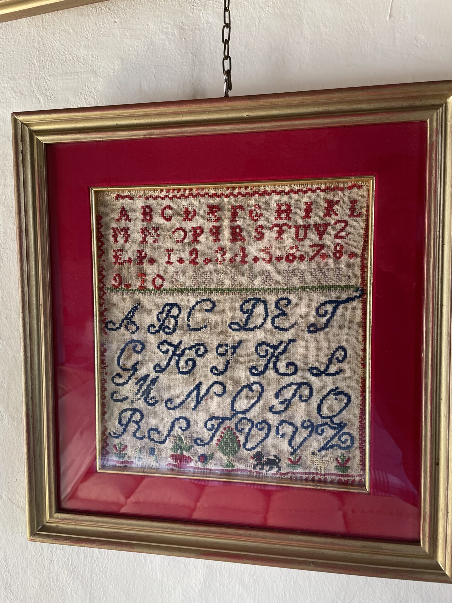 Piece of embroidery in a frame. It is an alphabet in red and blue. Technique used is cross stitch.